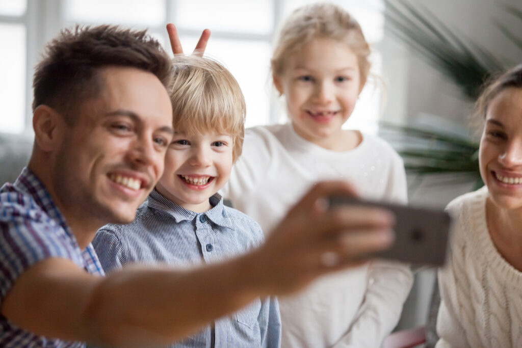 Kid sister making little cute brother bunny ears while father taking happy family selfie, funny children boy and girl having fun posing for self-portrait with smiling mom and dad bonding together