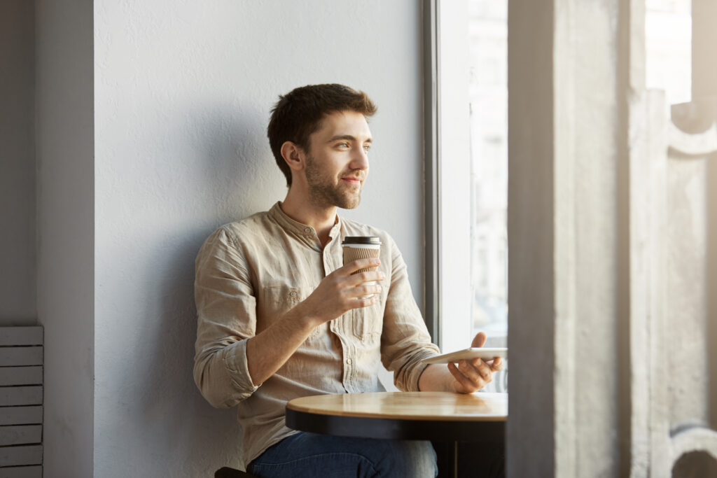 Portrait of young perspective male freelance designer sitting in cafeteria, looking aside being satisfied with his new project