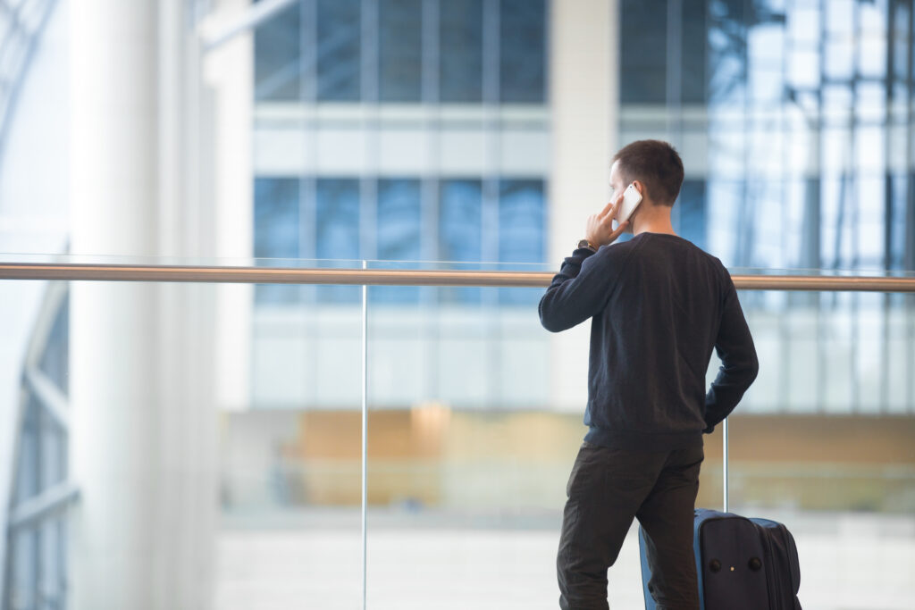 Young man in 20s talking on smart phone in airport, traveler standing beside luggage bag, wearing casual style clothes, rear view, copy space